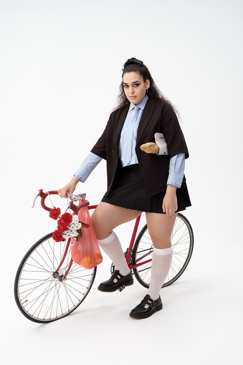 A person with long hair wearing a black jacket, blue shirt, and skirt stands next to a red bicycle adorned with flowers and a bag of groceries.
