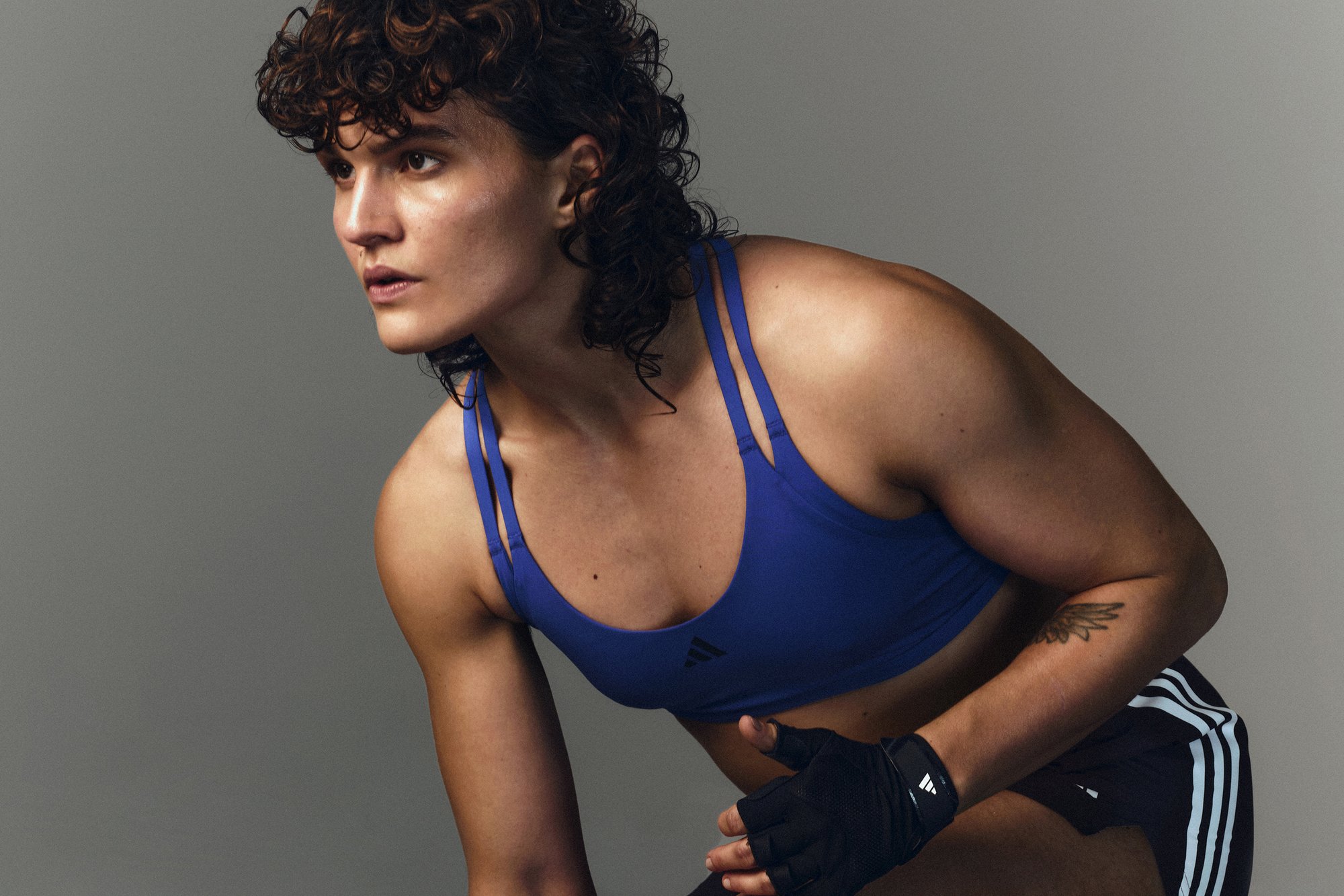 Athlete with curly hair in a blue sports bra and black gloves, poised in an athletic stance against a neutral grey background.