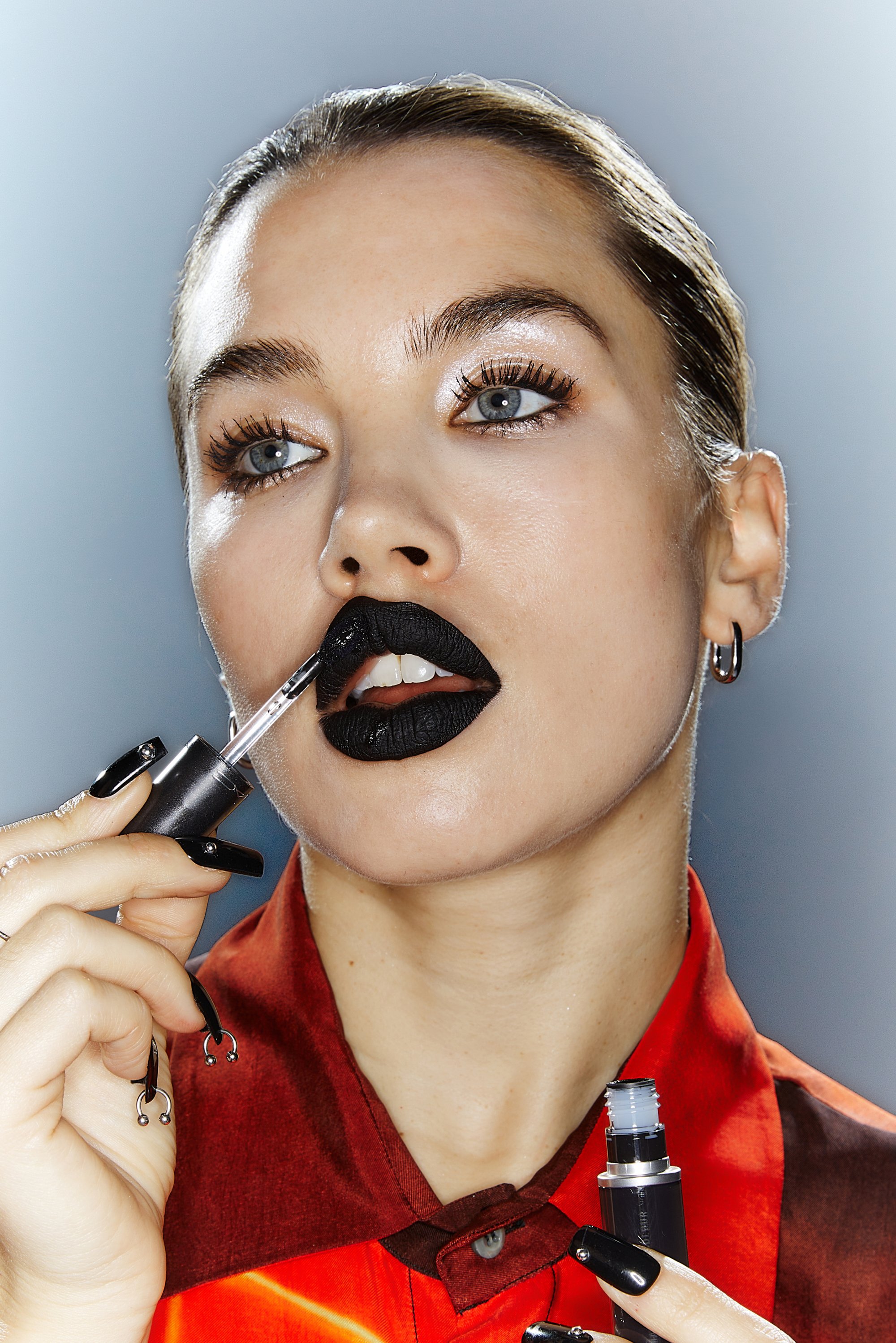 Model brengt een zwarte lipproduct aan met een applicator, gekleed in een rode shirt. Gedurfde make-up met dramatische wimpers en lange, zwarte nagels.