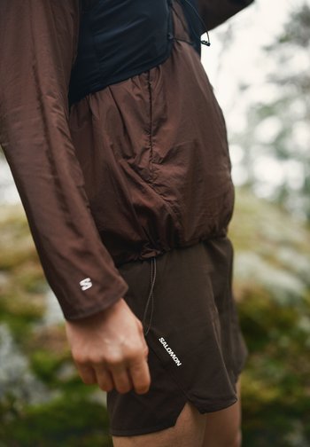 Sideprofil av en person som har på seg brune Salomon-shorts og en brun langermet jakke utendørs med uskarpt naturlig bakgrunn.