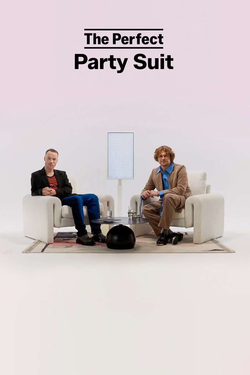 Twee mannen zitten op zachte fauteuils, de één in een zwarte blazer en blauwe broek, de ander in een beige pak, met een moderne tafel en decor.