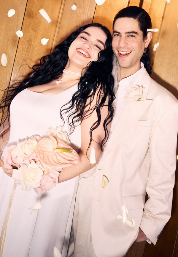 Abito da sposa bianco con una silhouette fluida, che tiene un mazzo di rose rosa chiaro e un dettaglio a forma di fenicottero. Completo beige con dettagli floreali.