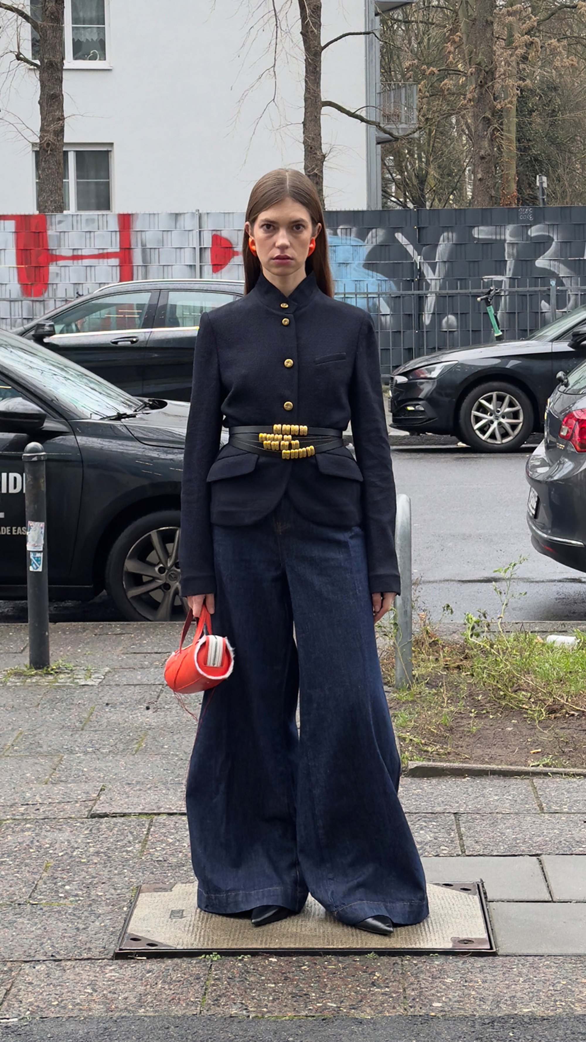 Una modelo está de pie en una acera, vistiendo una chaqueta negra a medida, jeans de pierna ancha y accesorios rojos, con grafitis y coches aparcados de fondo.