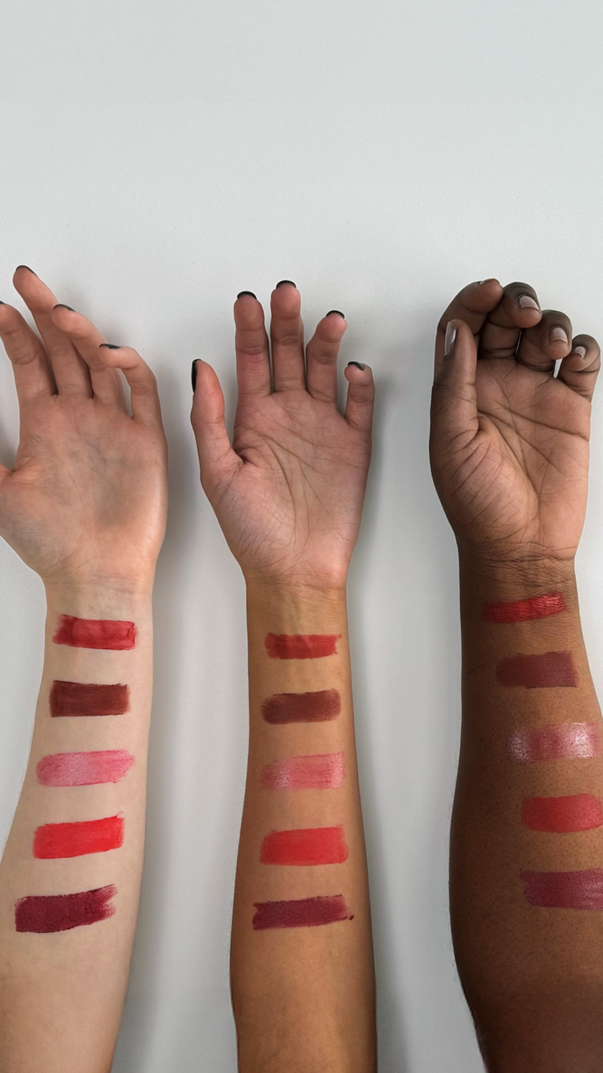 Tre braccia mostrano diverse tracce di rossetto in tonalità rosse su uno sfondo chiaro, evidenziando diverse tonalità di pelle e finiture.