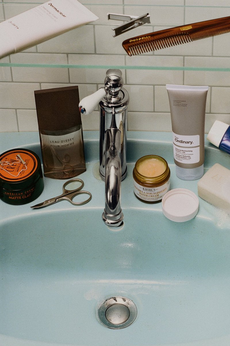 Waschbecken im Badezimmer mit silbernem Wasserhahn, umgeben von Pflegeutensilien wie Kamm, Schere, Hautpflegegläsern, Tube, Seife und Kölnflasche.