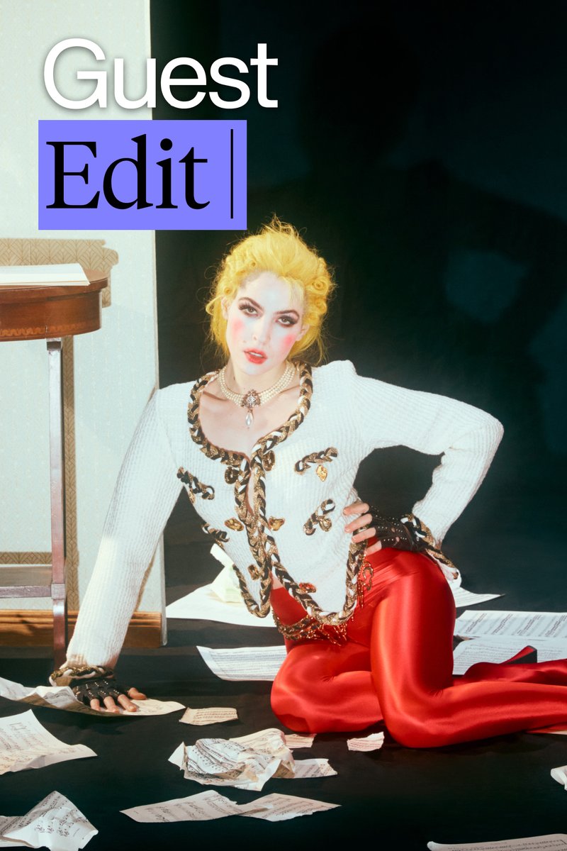 A person with yellow hair kneels amidst scattered papers, wearing a stylish white jacket with gold embellishments and red pants.