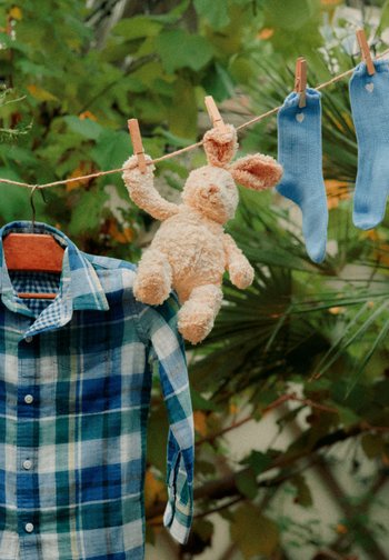 Blau kariertes Hemd auf Bügel, beiger Plüschhase und zwei blaue Babysocken hängen an einer Wäscheleine im Freien vor grünem Laub.