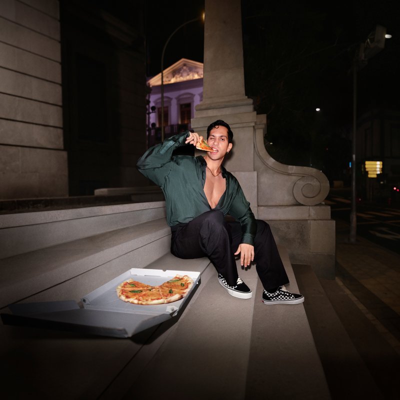Individuo sentado en escalones de piedra, vistiendo una camisa verde y pantalones negros, sosteniendo una porción de pizza por encima de una caja de pizza con una pizza parcialmente visible.