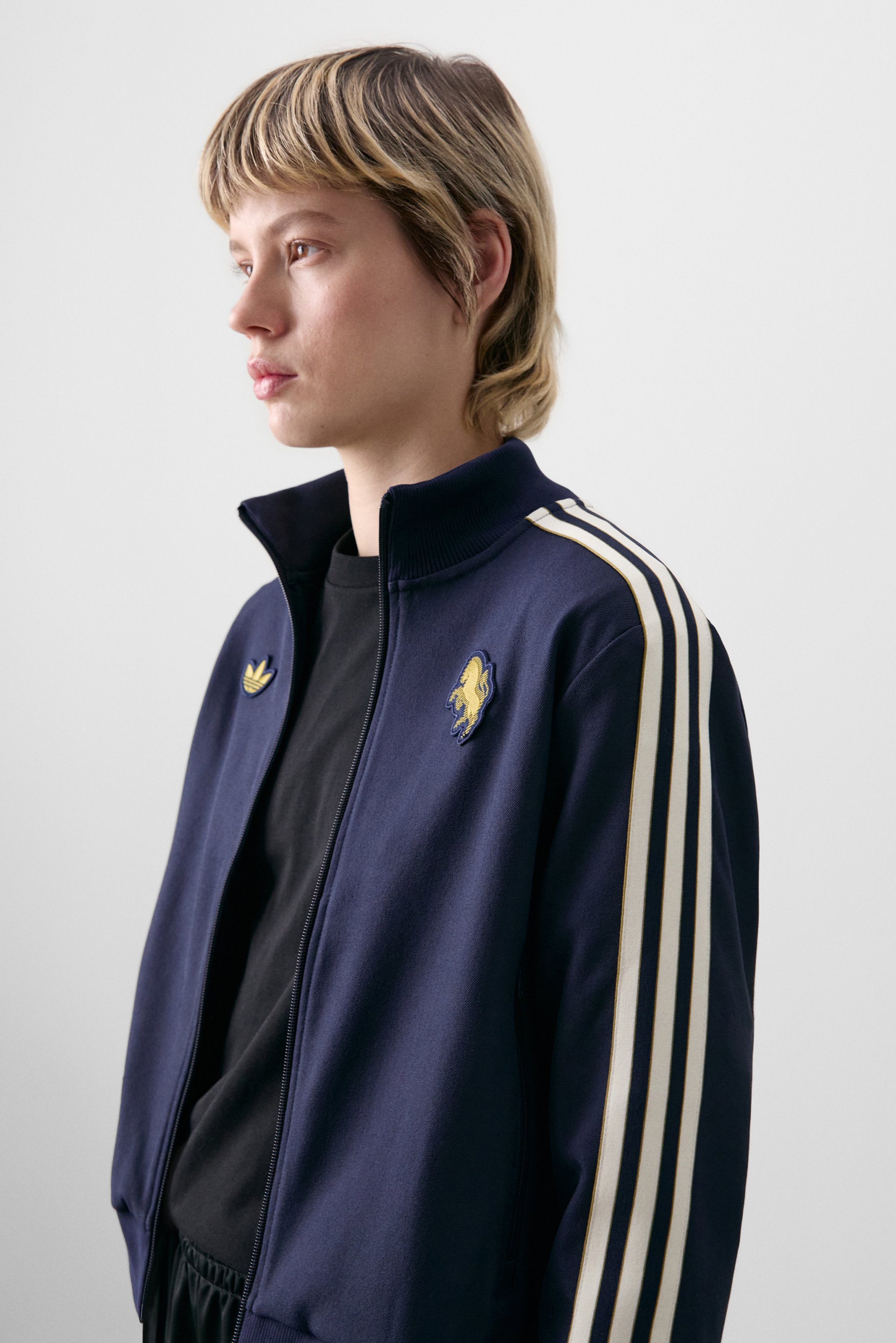 Persoon met kort blond haar die een marineblauwe hoodie met rits draagt, voorzien van witte strepen en een logo, tegen een neutrale achtergrond.