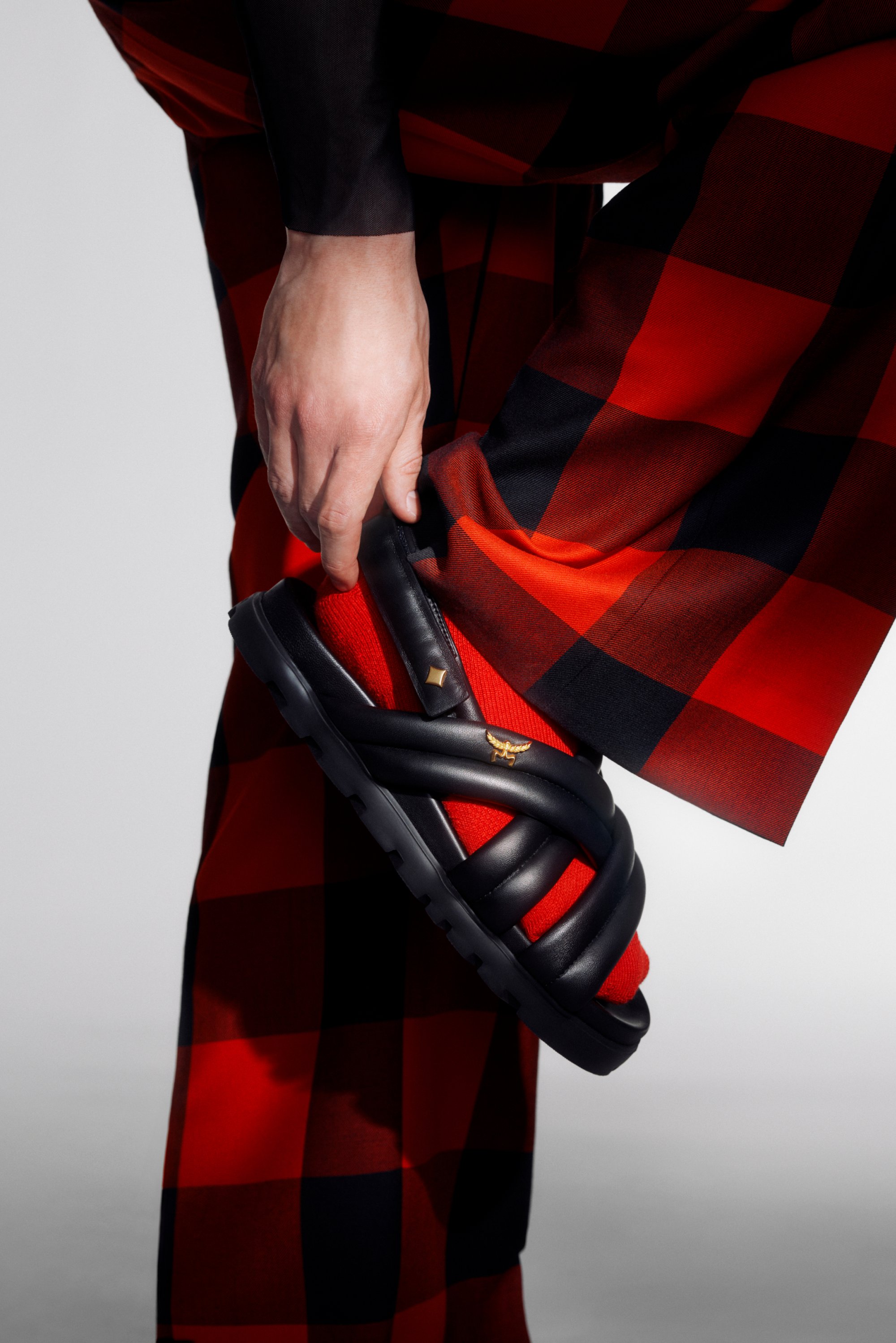 Una mano ajusta una sandalia negra con tiras, que se lleva con pantalones de cuadros rojos y negros, destacando la moda urbana elegante.