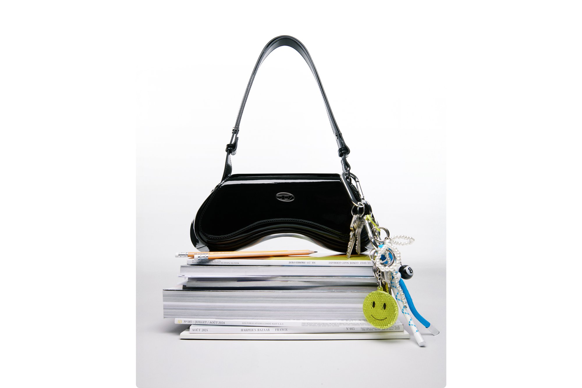 A black handbag rests on a stack of magazines, accompanied by a pencil and keychain with multiple colorful charms, including a smiley face.
