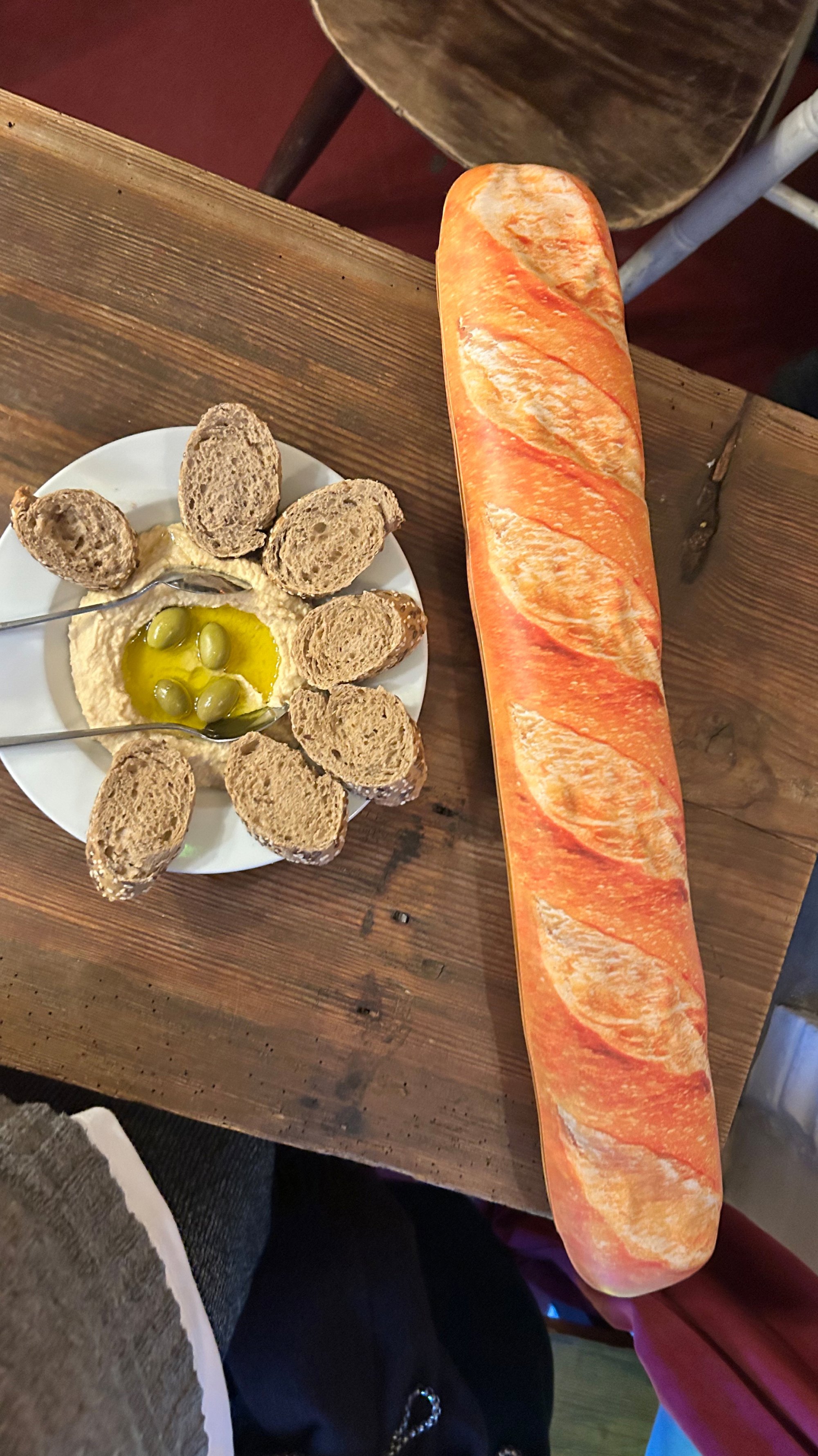Een lange baguette ligt naast een bord hummus, omringd door kleine bruine broodstukjes en groene olijven, op een houten tafel.