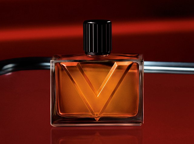 Flacon de parfum rectangulaire en verre ambré avec un bouchon noir côtelé et un grand V en relief sur le devant, sur fond rouge foncé.