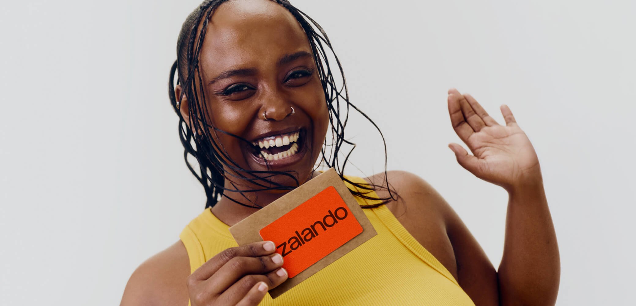Lächelnde Frau in gelbem Top hält eine braune Karte mit einem orangefarbenen Zalando-Logo und winkt mit der anderen Hand vor weißem Hintergrund.