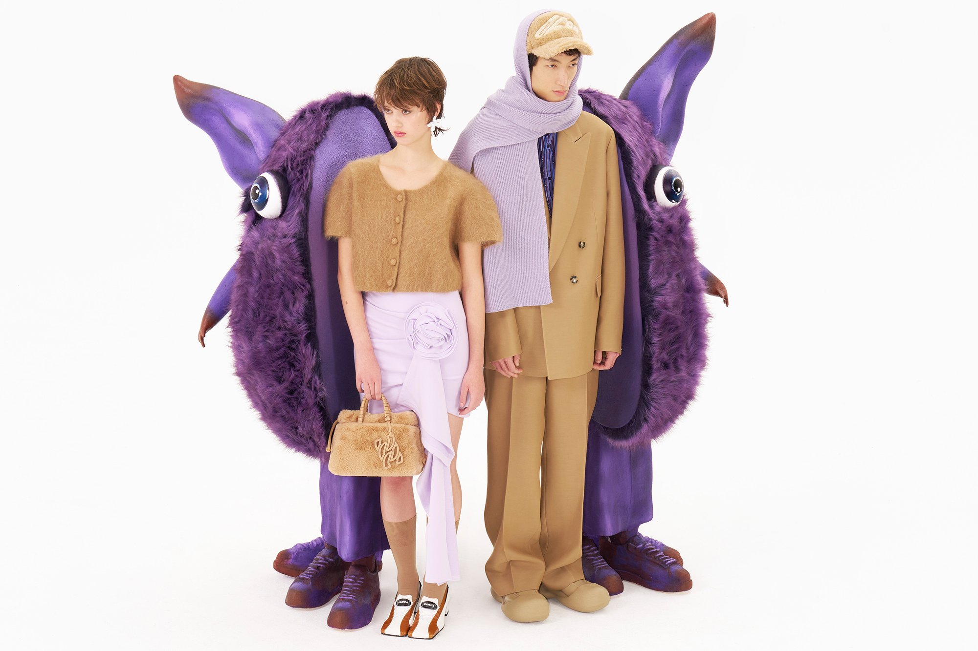 Two stylish models stand together, flanked by large purple furry creatures with prominent eyes, against a plain white background.
