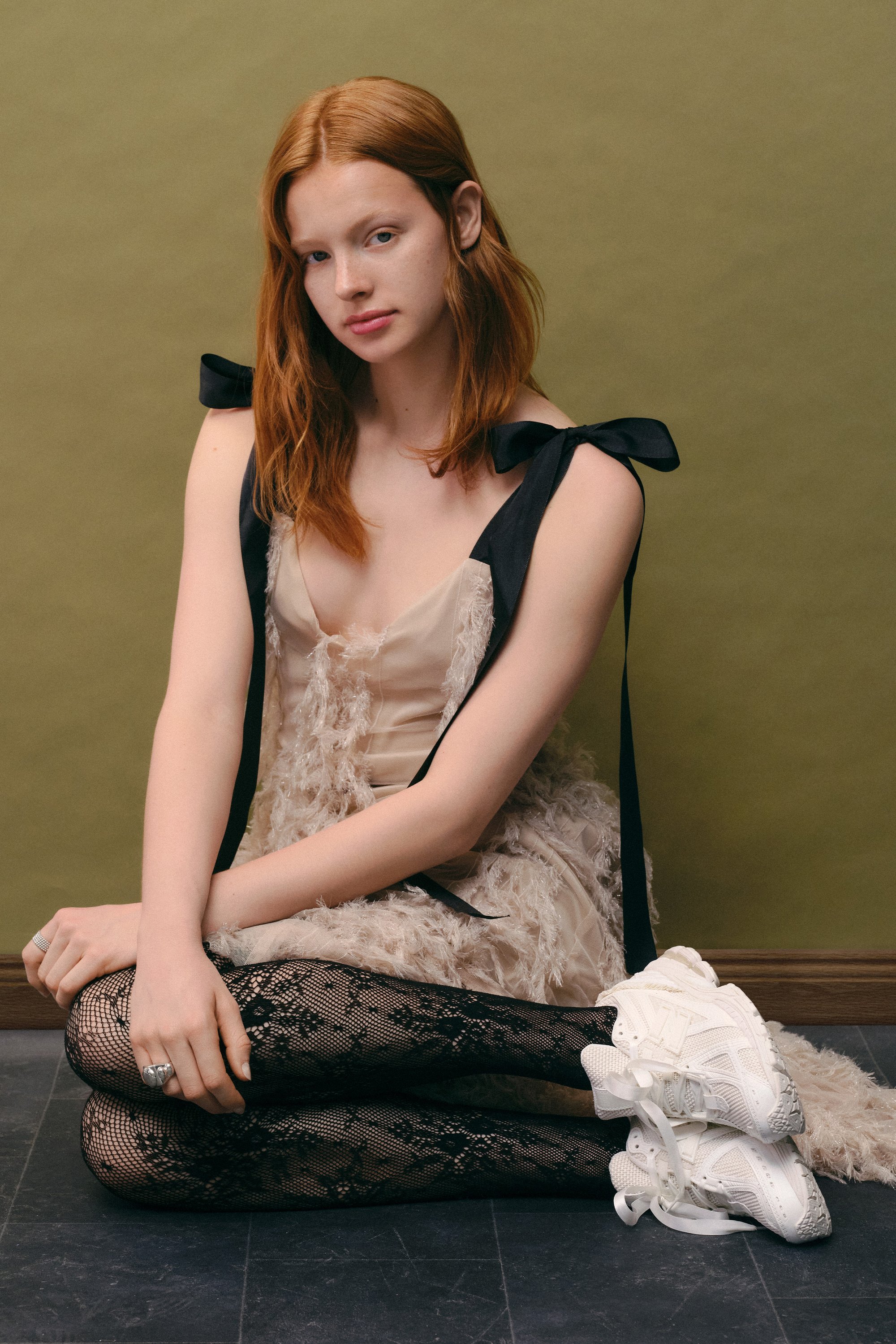 A young woman with red hair sits cross-legged, wearing a cream dress with textured layers, lace tights, and white sneakers, against a green backdrop.