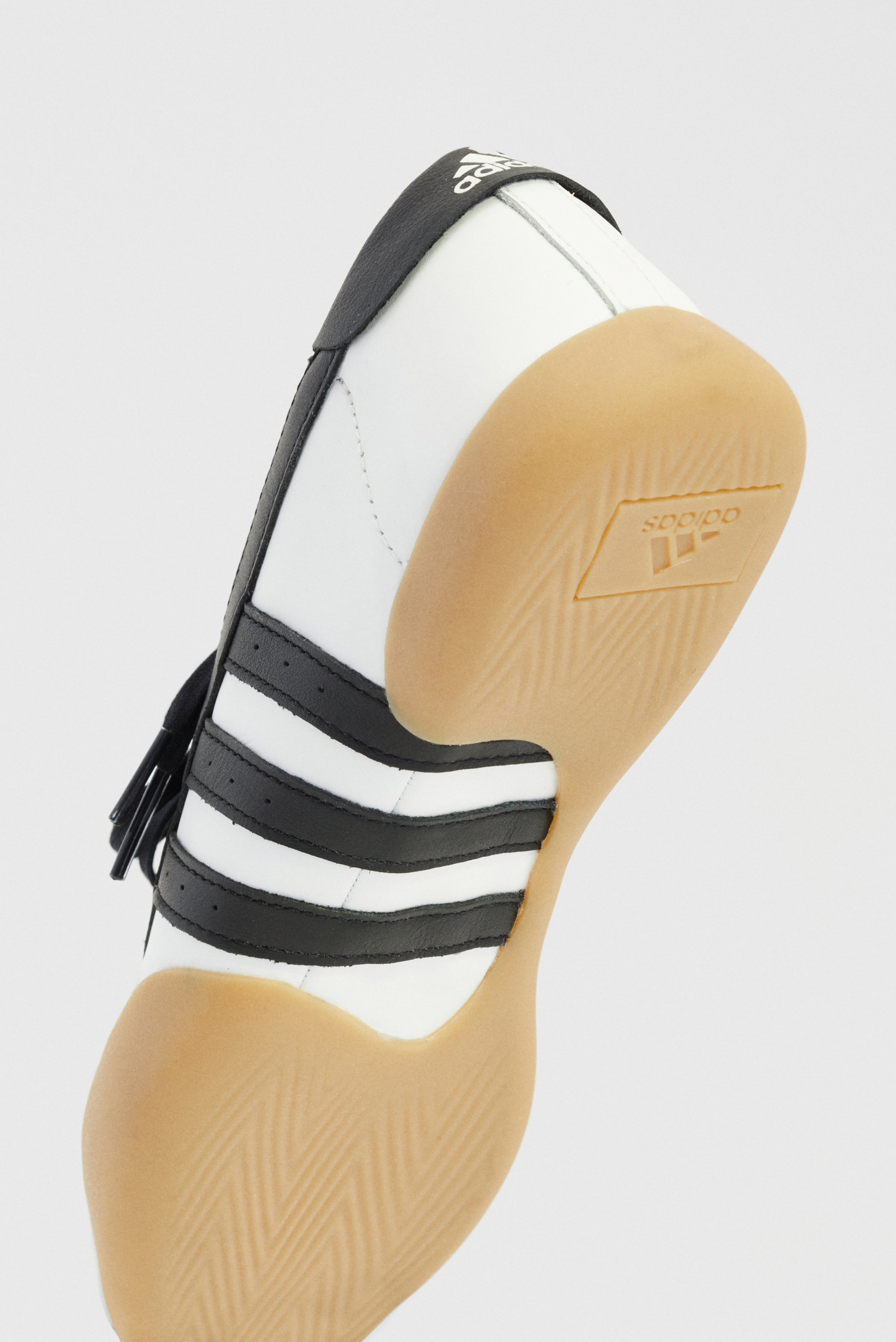 White leather trainers with black stripes, rounded toe, rubber gum sole, and a visible logo on the tongue. They feature a smooth and textured design.
