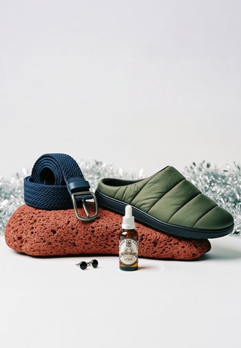 Grønn quiltet slip-on sko, vevd marineblå belte med metallspenne, ravfarget skjeggolje flaske, og små svarte runde solbriller på en rød vulkansk stein.