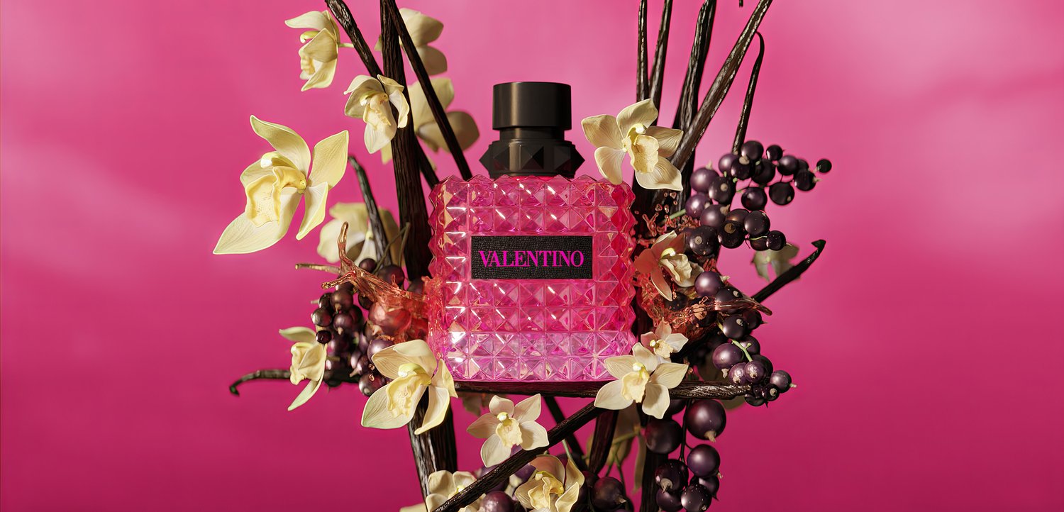 Pink Valentino perfume bottle surrounded by cream flowers and dark purple berries on dark branches against a bright pink background.
