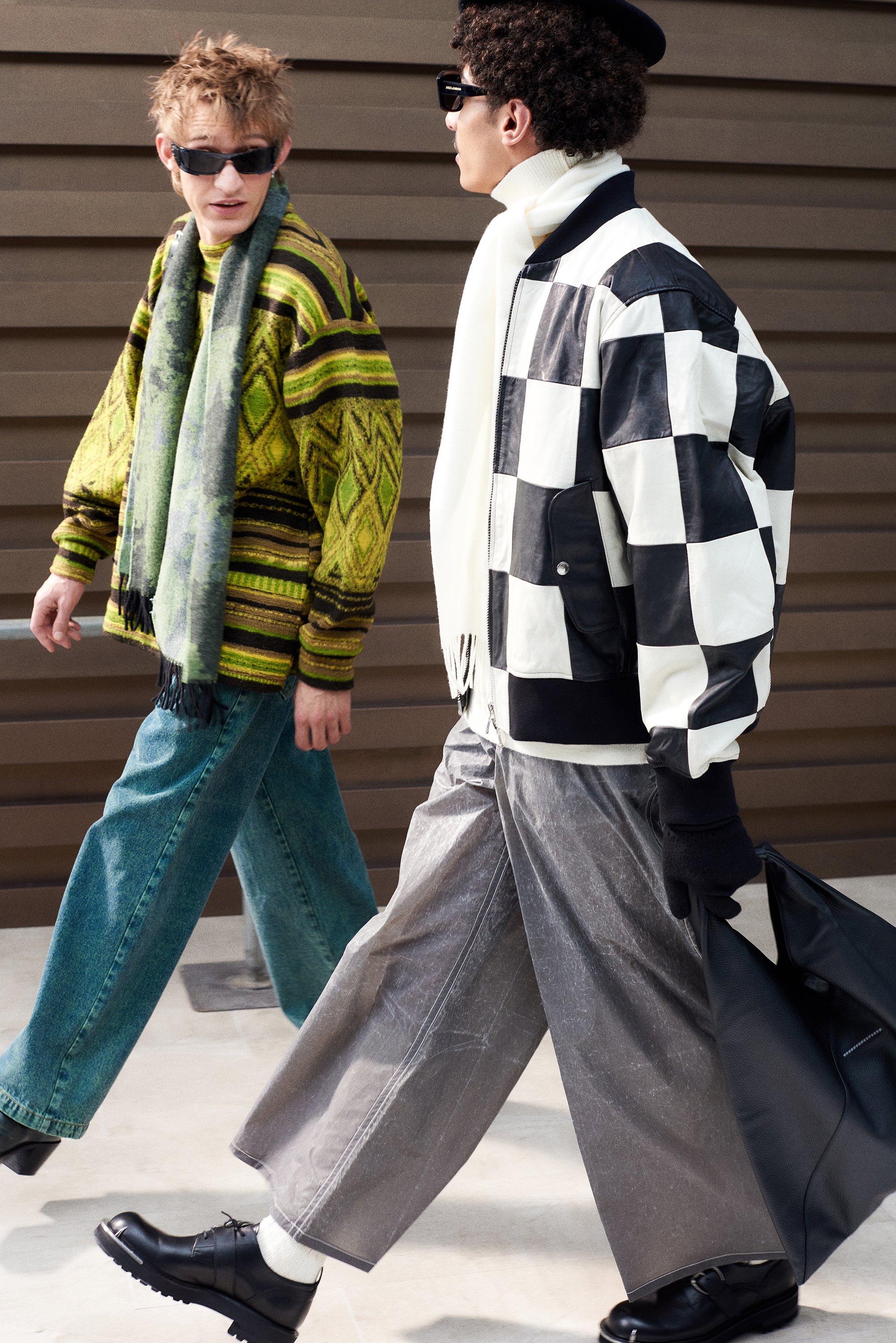 Two young men walk side by side, showcasing distinctive fashion: one in a patterned sweater and scarf, the other in a checkered bomber jacket.