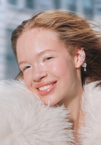 En kvinde med lys hud smiler, iført sølvøreringe formet som spiraler, indrammet af flydende brunt hår og en hvid pelskrave.