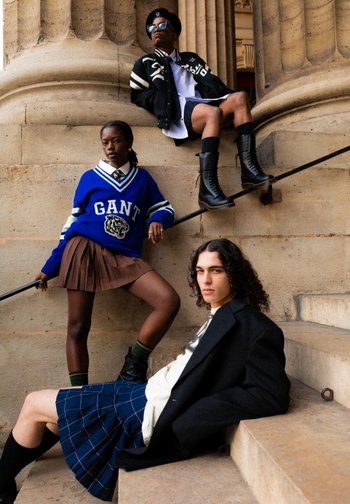 Drei junge Erwachsene posieren auf einer Treppe: einer in einem blauen Pullover, ein anderer in einer schwarzen Jacke und Shorts und eine dritte in einem schwarzen Rock und Blazer.