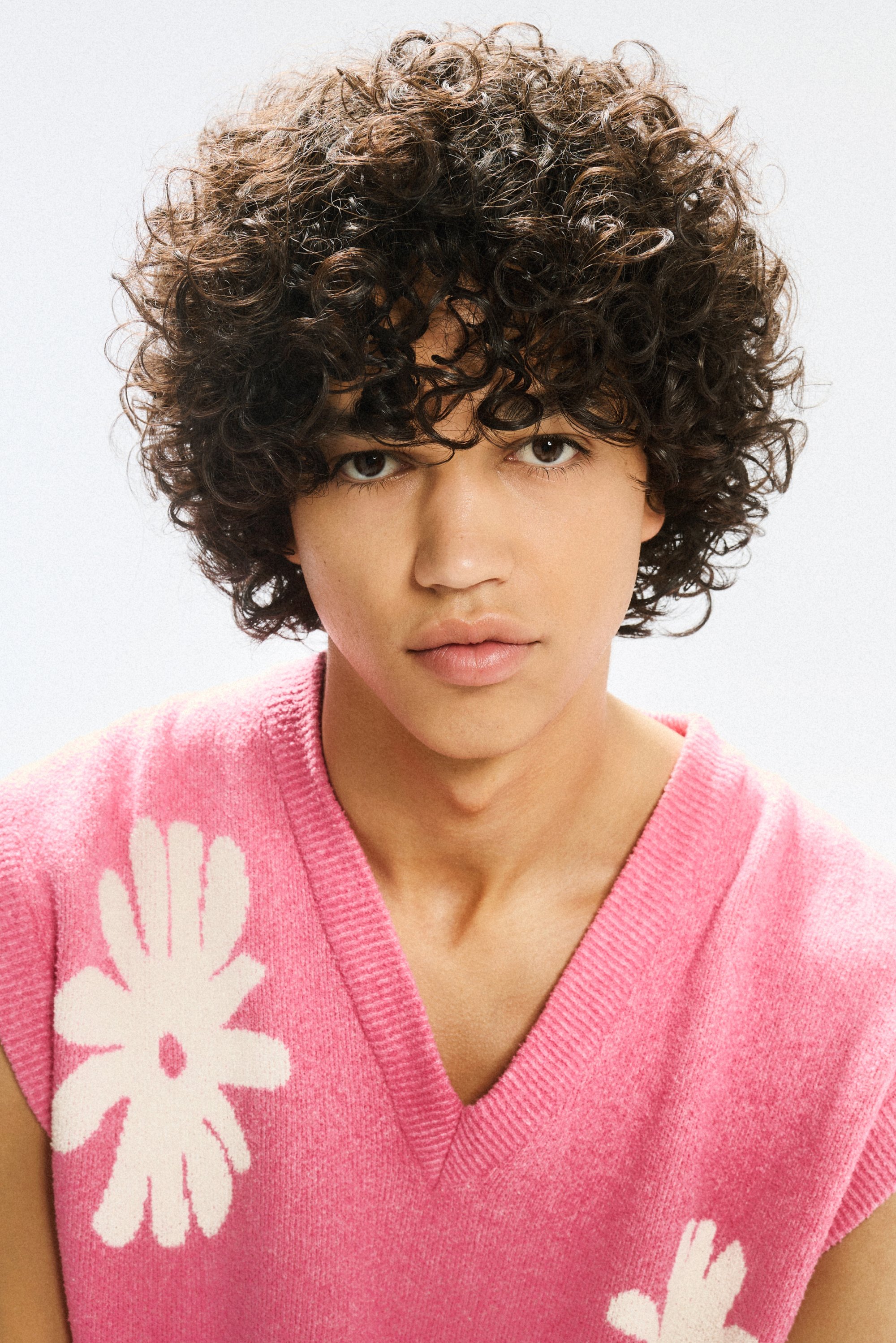 Young person with curly hair, wearing a pink sweater featuring a white floral design, gazes directly at the viewer against a light background.