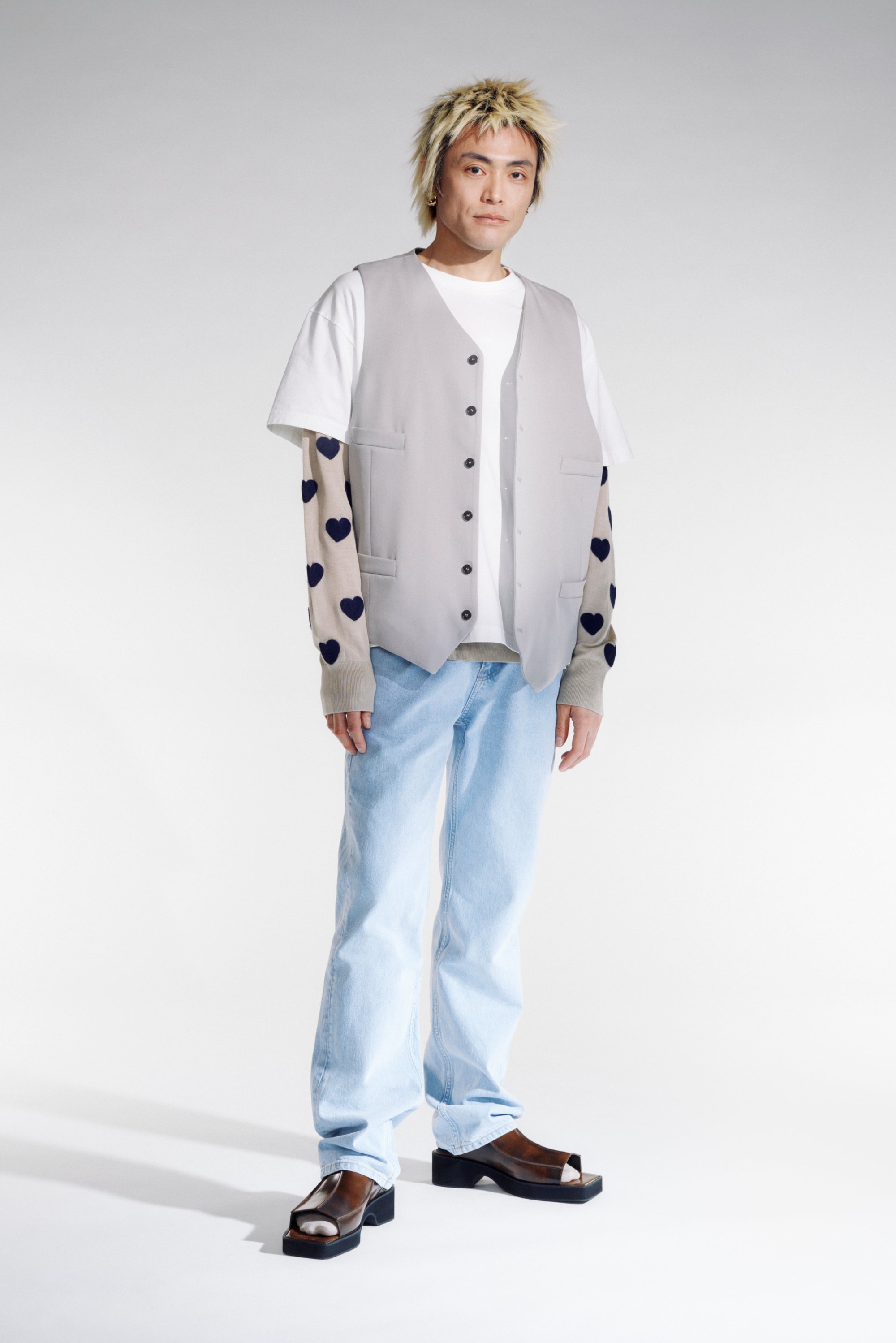 Een jong persoon staat tegen een lichte achtergrond, gekleed in een grijze vest over een wit shirt, lichte spijkerbroek en dikke sandalen.