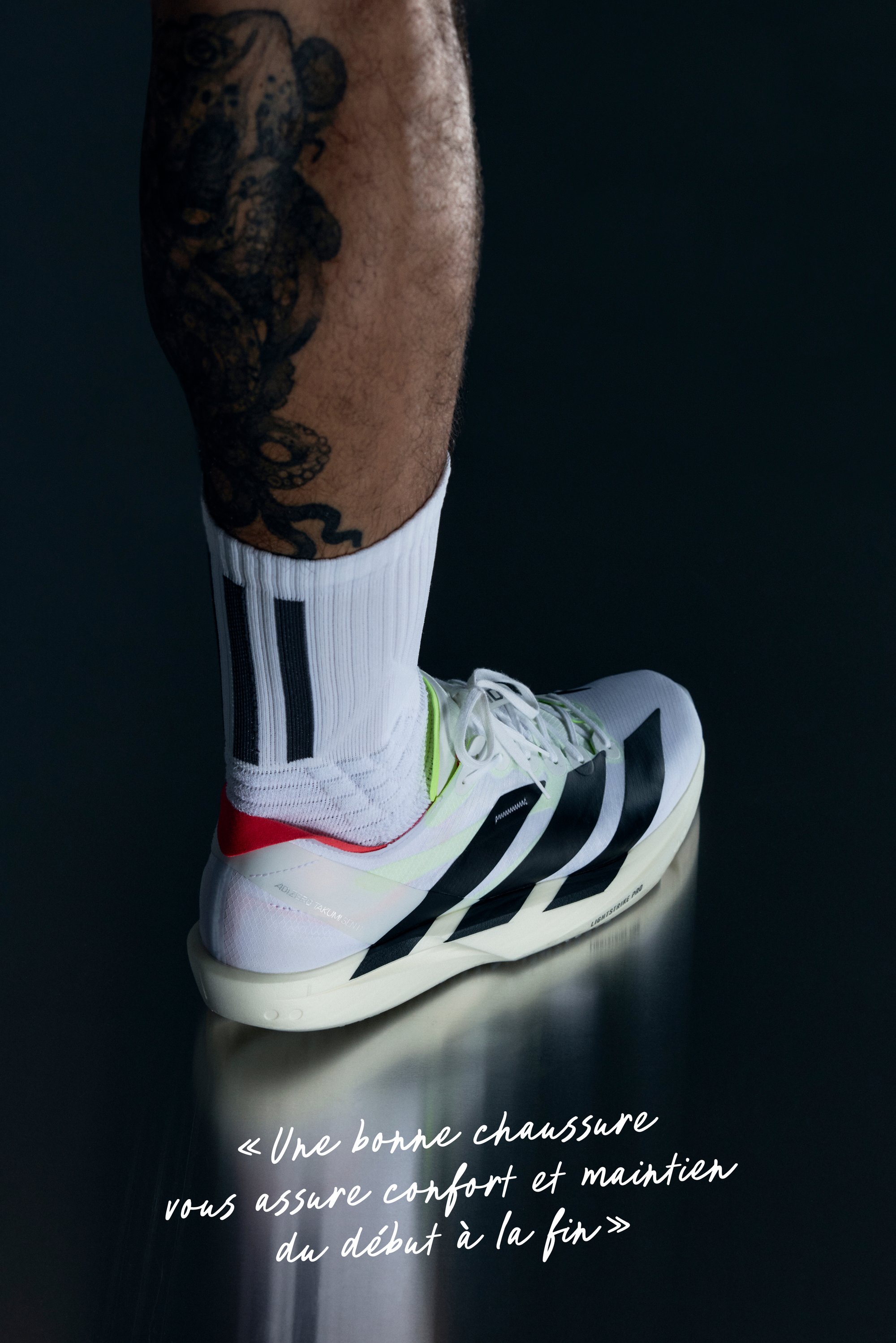 Jambe inférieure d'un athlète avec un tatouage de pieuvre, portant une chaussette Adidas blanche et une chaussure de course blanche avec des rayures noires sur une surface sombre réfléchissante.