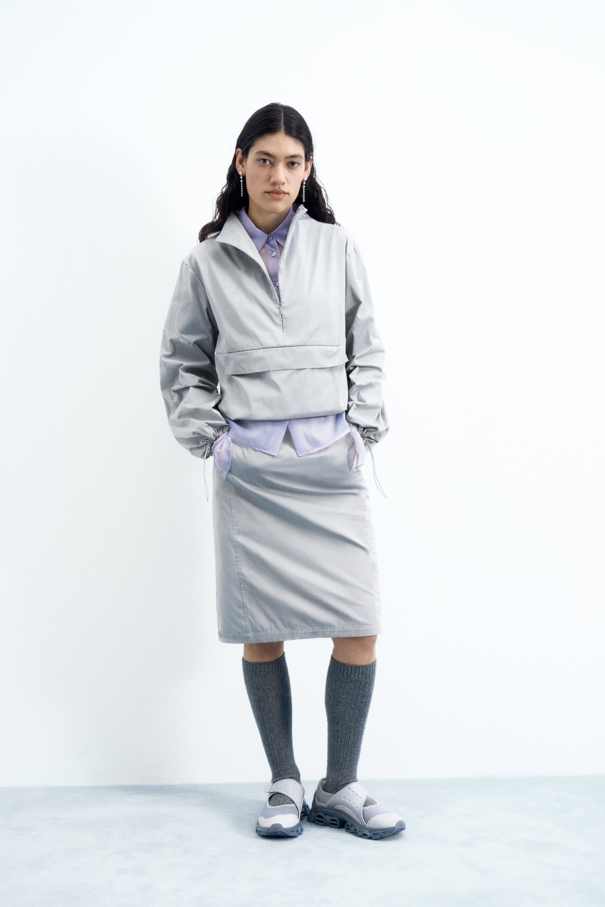 Una joven posa con una chaqueta gris claro y una falda a juego, combinadas con calcetas altas grises y zapatillas deportivas, contra un fondo blanco.