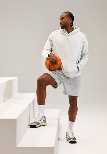 A person stands on a step, holding a basketball, wearing a gray hoodie and shorts, with a neutral background highlighting the outfit.