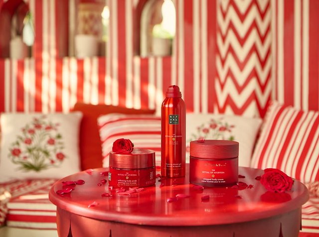 Rituel Rouge des produits de soin du corps Ayurveda sur une table ronde rouge avec des pétales de rose, sur un fond rayé rouge et blanc et floral.