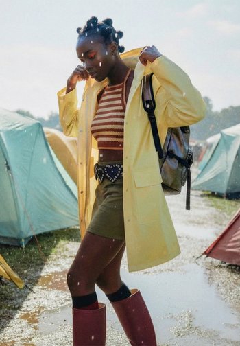 Junge Person richtet den gelben Regenmantel, während sie in einer matschigen Pfütze auf dem Campingplatz steht, mit gestreiftem Oberteil, Shorts, Gummistiefeln und Rucksack.