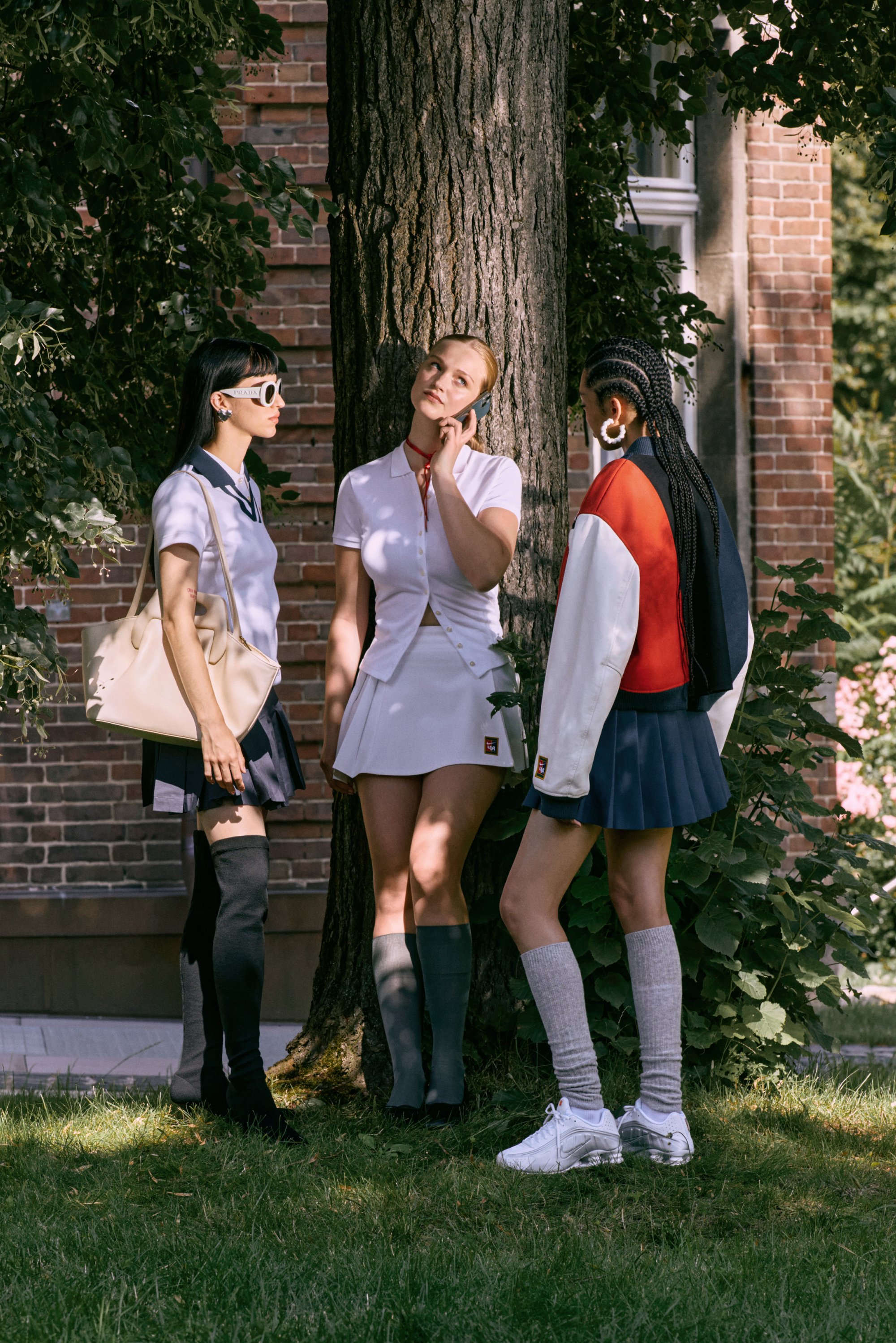 Three women stand by a tree, wearing stylish outfits, engaged in conversation. One is on her phone, surrounded by greenery and a brick building.