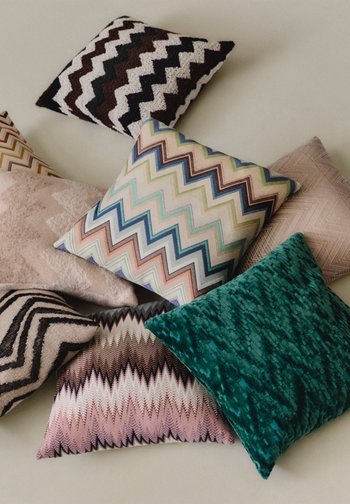 Οκτώ διακοσμητικά μαξιλάρια με διάφορα ζιγκ ζαγκ και υφασμάτινα σχέδια σε παστέλ, καφέ, μαύρους και τιρκουάζ τόνους πάνω σε ανοιχτή επιφάνεια.