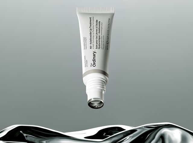 Un tube blanc de soin des lèvres The Ordinary HA flotte au-dessus d’une surface liquide brillante en noir et blanc, sur fond gris.