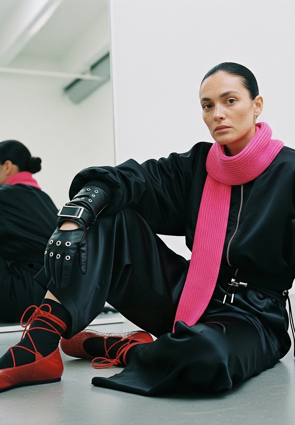 A woman with slicked-back hair is sitting on the floor, dressed in a black outfit, a bright pink scarf, black leather gloves, and red lace-up shoes, with her reflection visible in the mirror.
