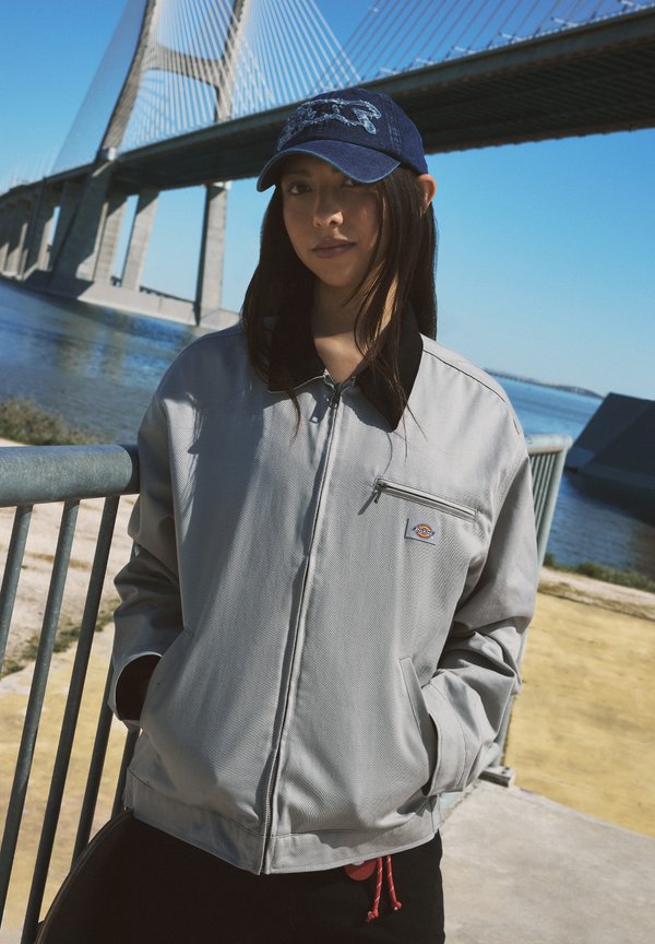 Young woman wearing light grey jacket and blue cap stands by waterfront railing with large cable-stayed bridge in background under clear sky.