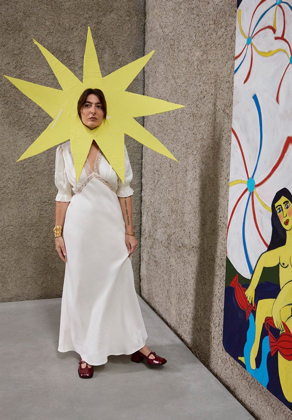 Witte satijnen jurk met kanten accenten, oversized geel papieren sterrenhoofdband, en bordeauxrode schoenen met blokhak, tegen een getextureerde muur en schilderij.