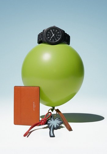 Black wristwatch resting on a green balloon, which is tied to a leather cardholder, a red cord, and a palm tree keychain on a white surface with gentle shadows.