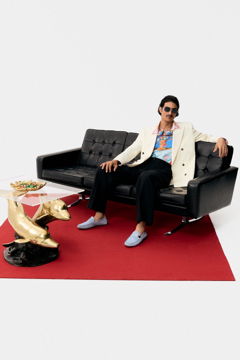 Ein Mann in einem weißen Blazer und Sonnenbrille entspannt sich auf einem schwarzen Sofa, mit einer goldenen Delfinskulptur und einem Snacktisch auf einem roten Teppich.