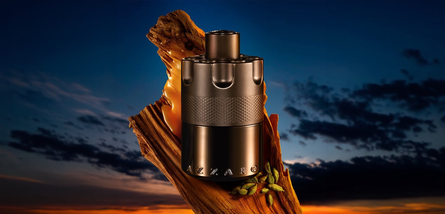 Flacon de parfum métallique Azzaro posé sur un morceau de bois texturé avec des capsules de cardamome, sur fond de ciel dramatique au crépuscule.