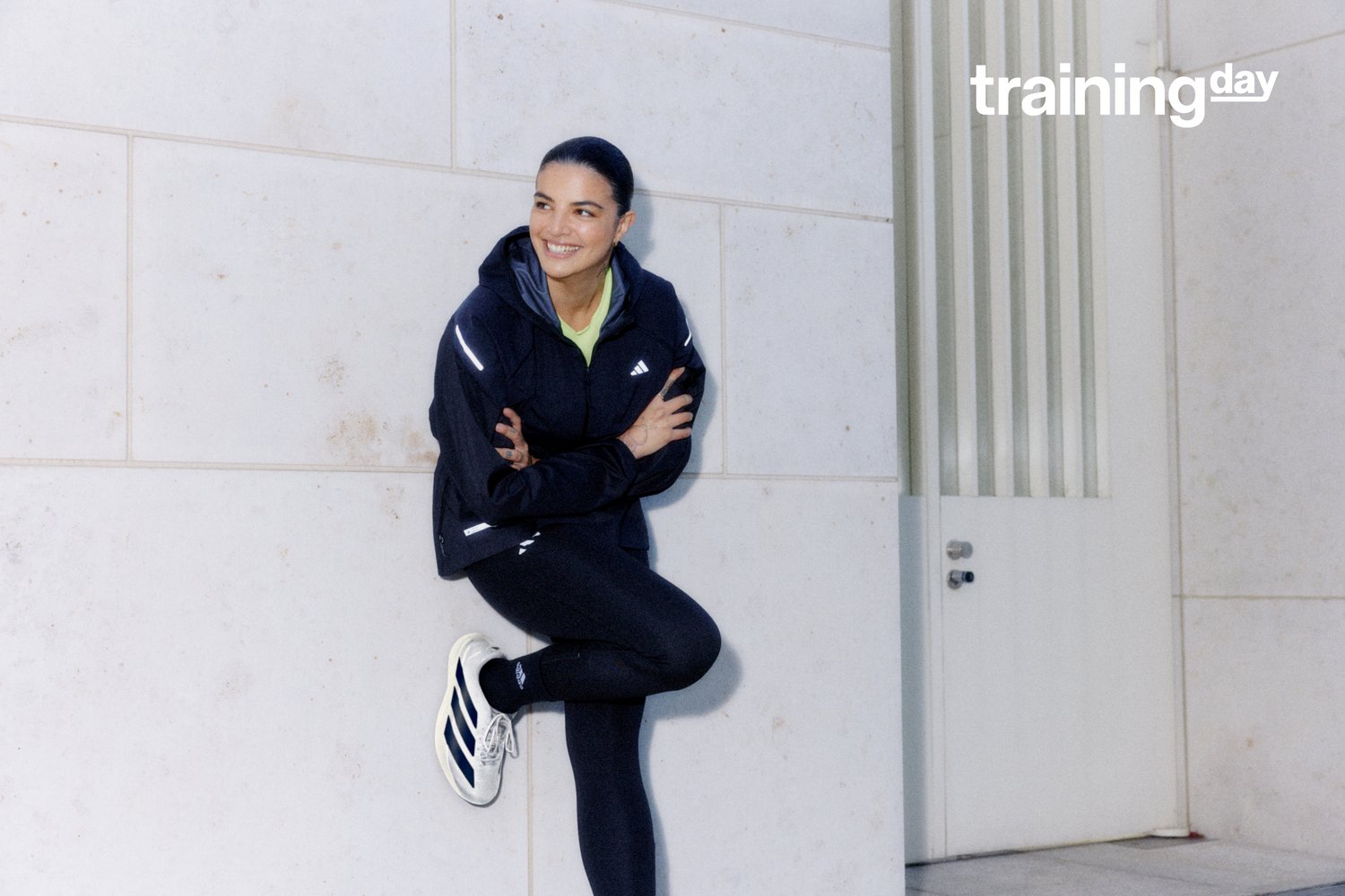 Una mujer sonriente vestida con ropa deportiva se apoya contra una pared de color claro, con los brazos cruzados. En el fondo se puede ver una puerta y franjas verticales.