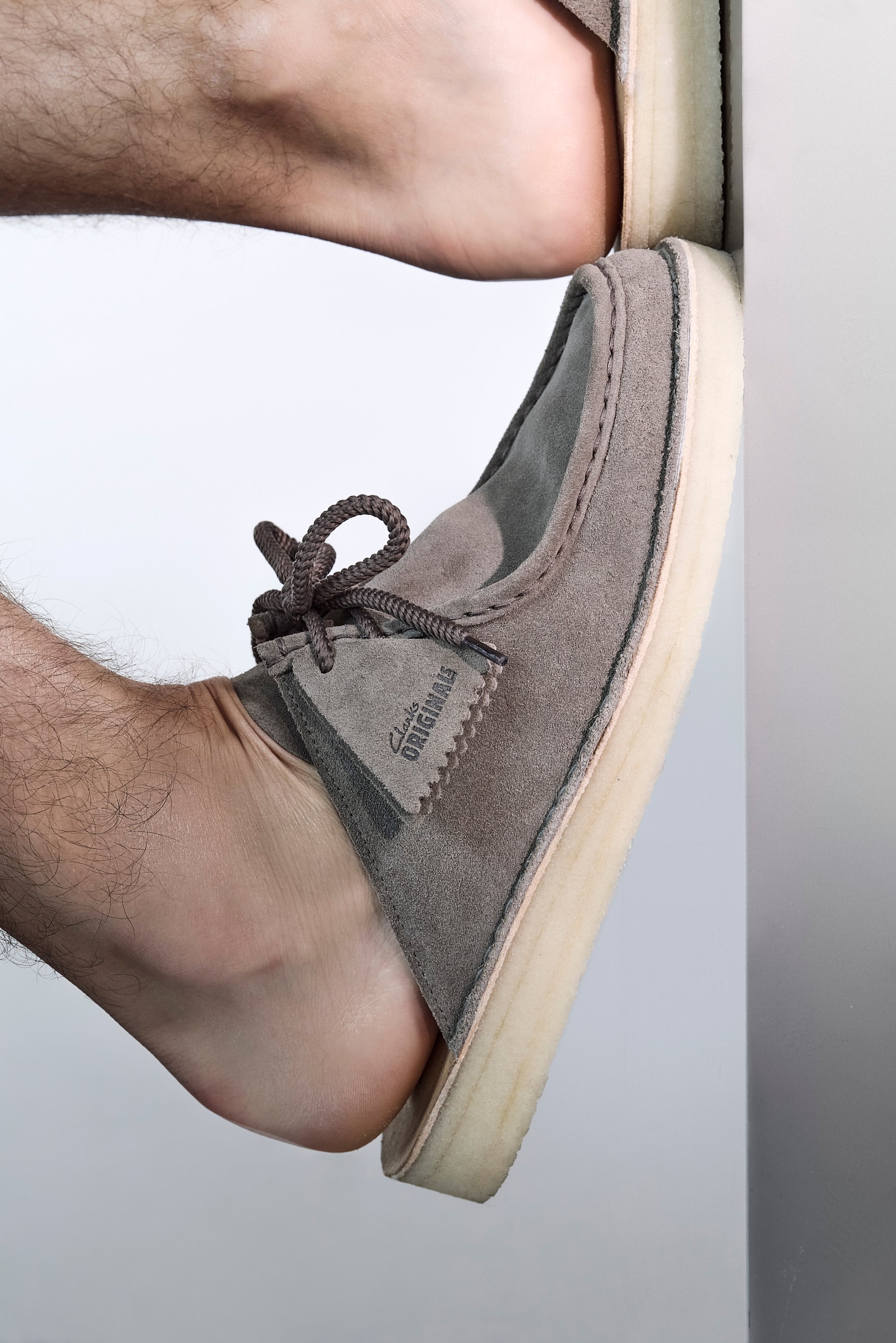 A foot clad in a grey suede shoe is placed against a vertical surface, highlighting the shoe's detail and texture.