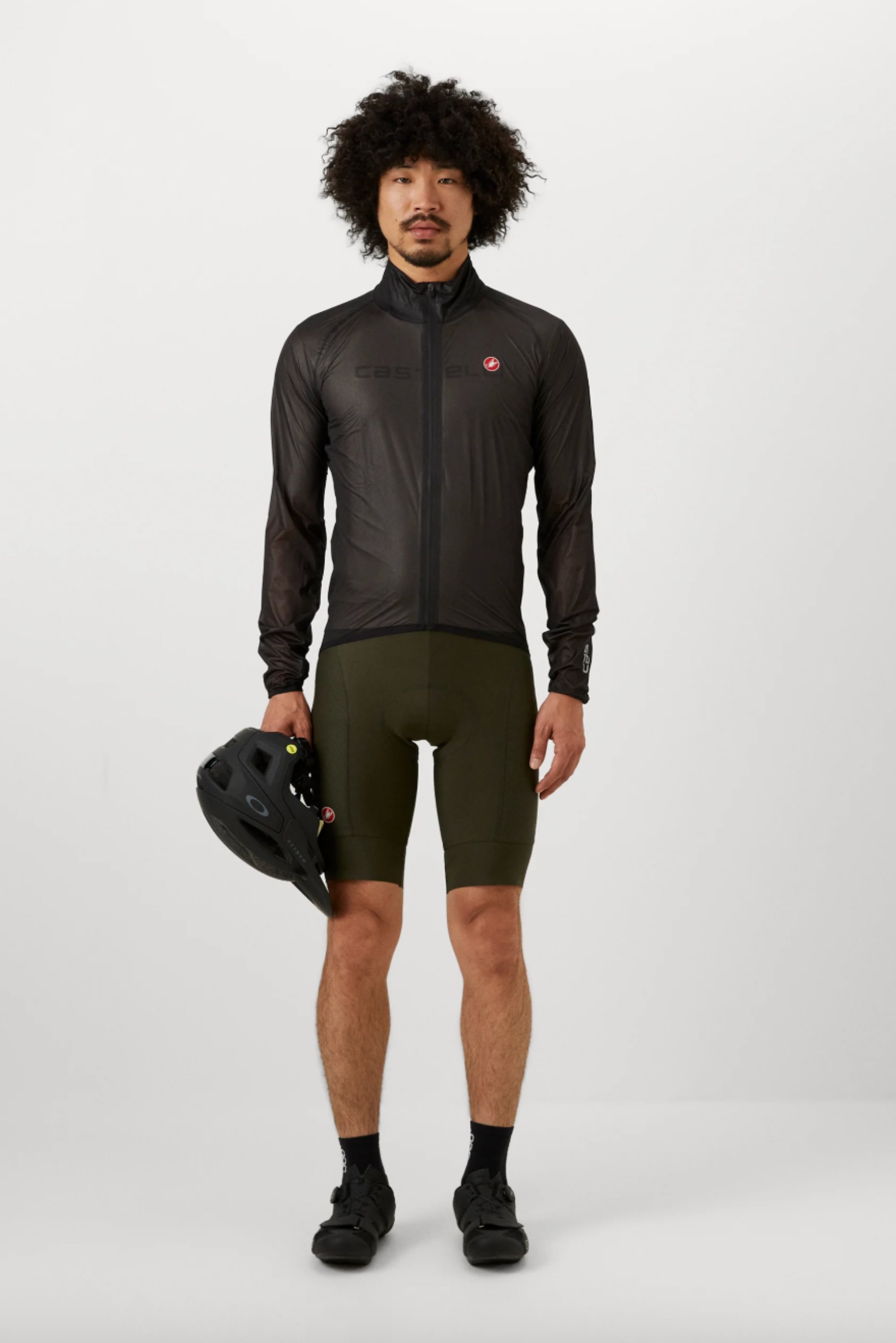 Ein Mann mit lockigem Haar steht in einer schwarzen Radjacke und olivgrünen Shorts, wobei er einen schwarzen Helm in einer Hand hält.