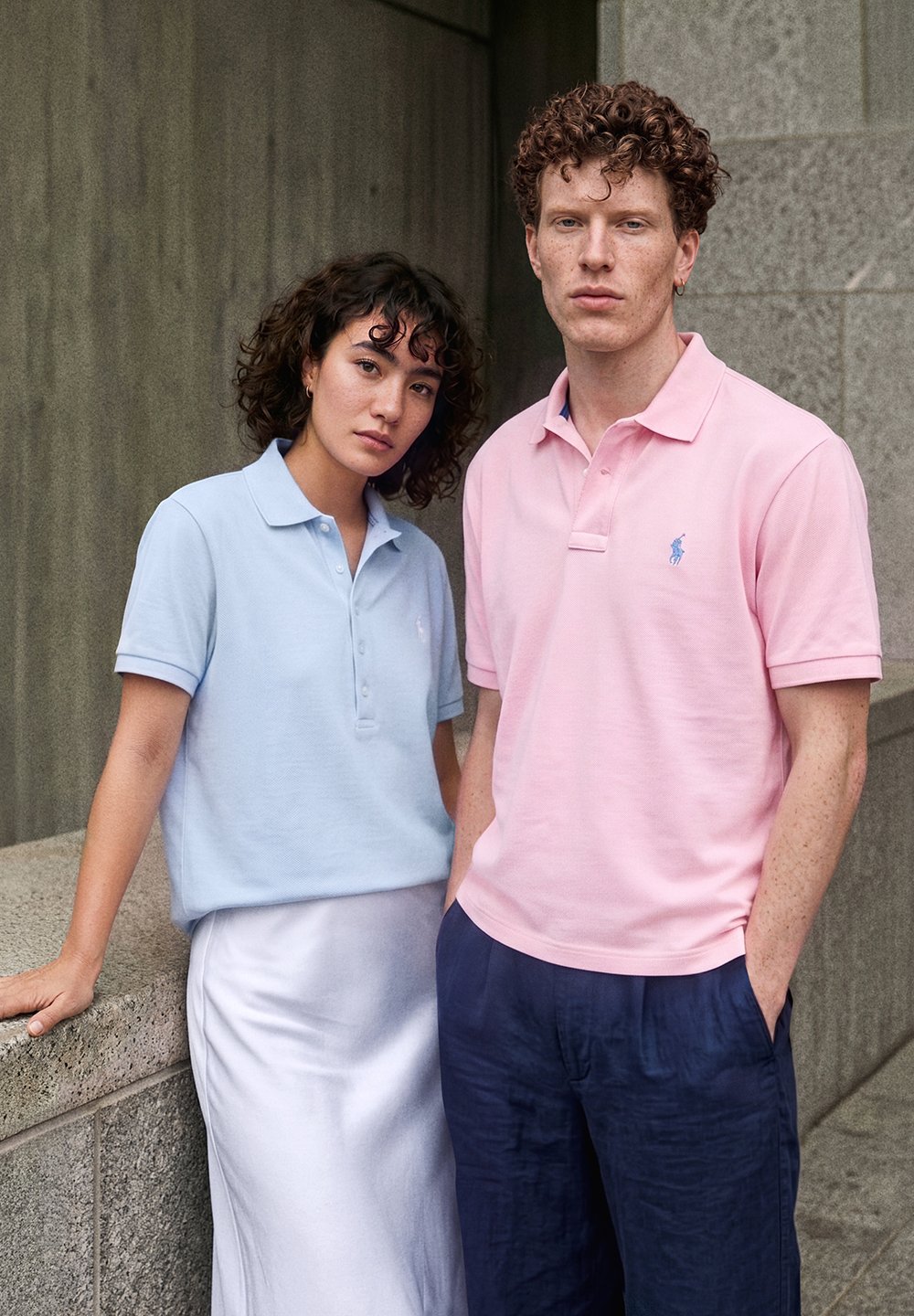Kvinne i lys blå polo og hvit skjørt som lener seg på en steinkant, mann i rosa polo og marineblå bukser står ved siden av henne mot en grå vegg.