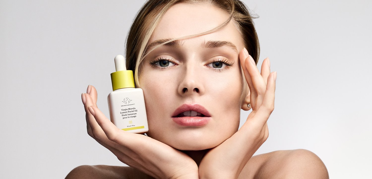 Woman with neutral expression holds a yellow-capped skincare serum bottle near her face, hands framing her cheeks against a plain background.