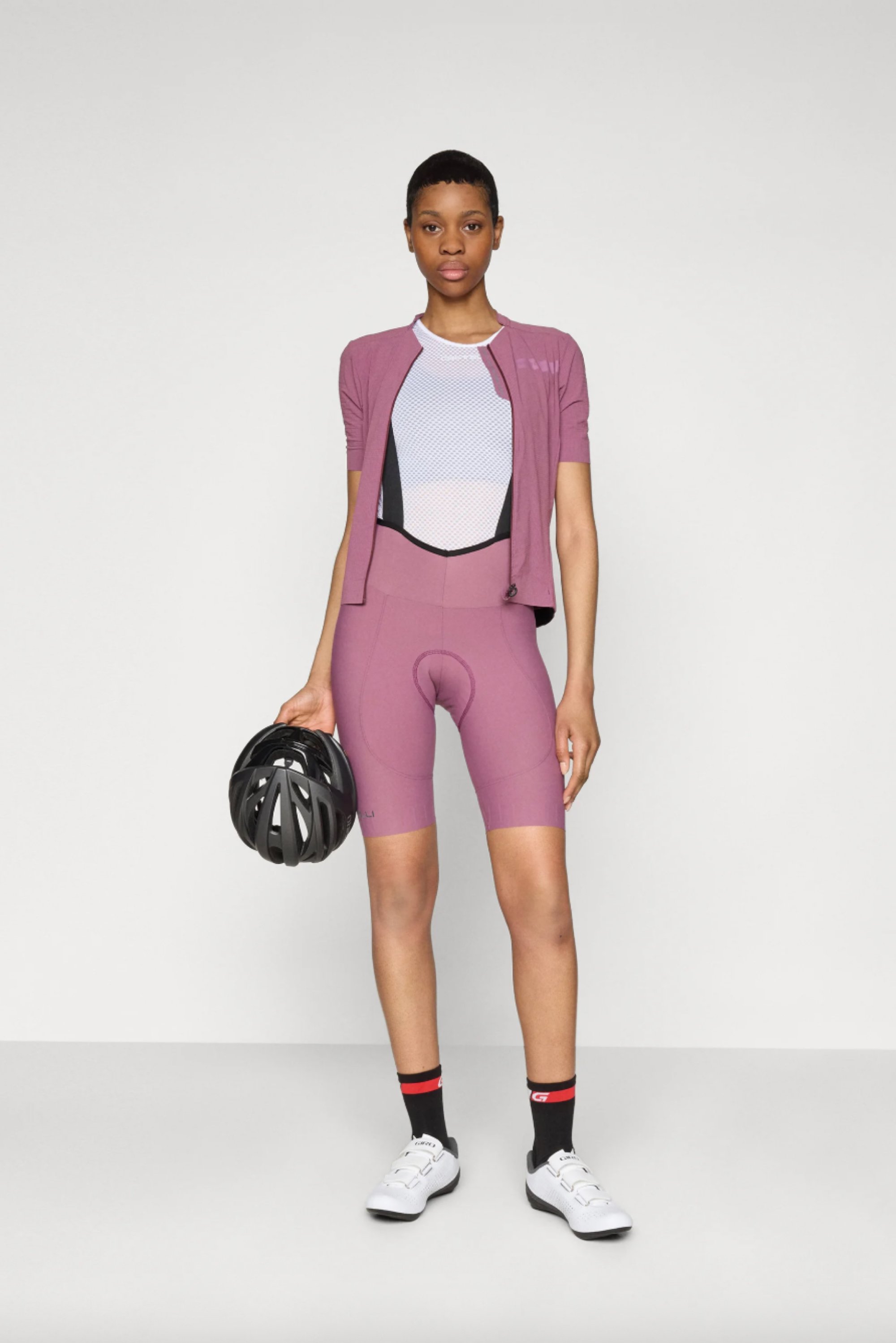 A person stands in sporty attire featuring a purple jersey and cycling shorts, holding a black helmet, against a plain background.