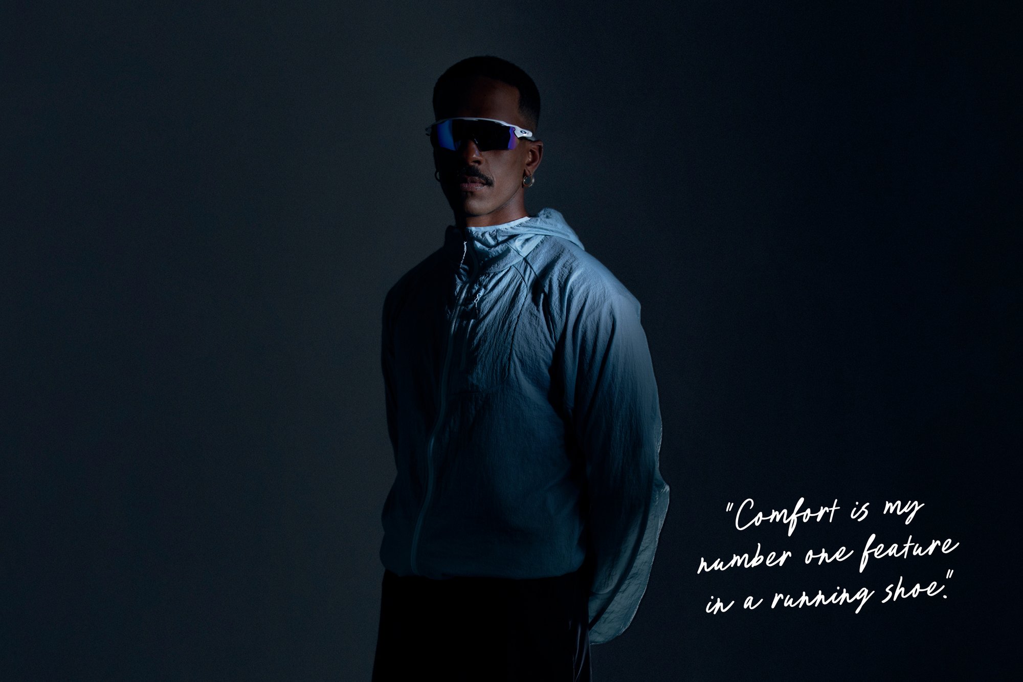 Helle blaue, leichte Laufjacke mit Kapuze, stehendes Modell im Profil. Sonnenbrille mit verspiegelten Gläsern und Zubehör, dunkler Hintergrund.