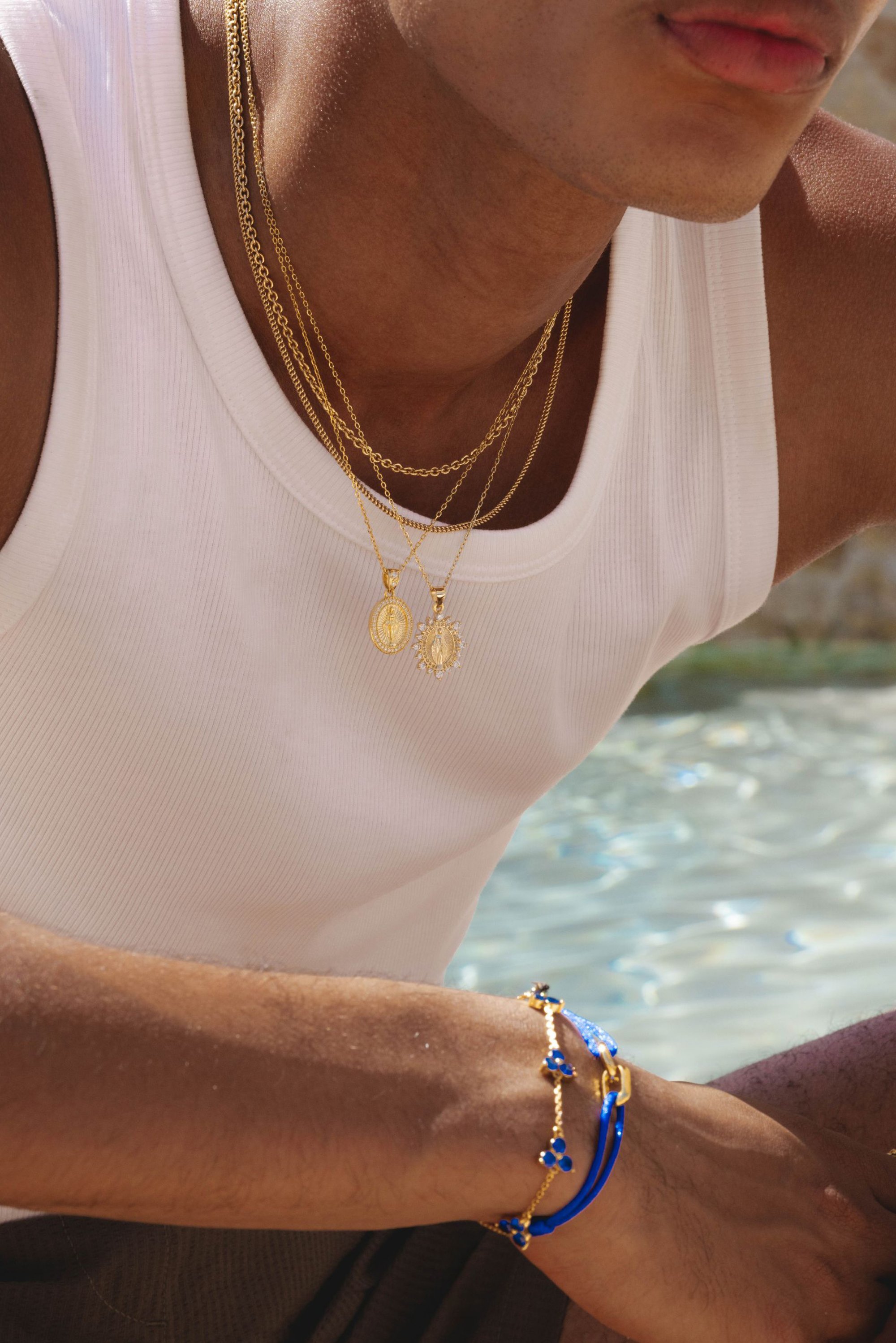 Collares de cadena de oro con colgantes, camiseta sin mangas blanca, pulsera con acentos azules y herrajes dorados. Textura de piel suave, fondo de piscina.